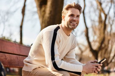 Neşeli bir adam güneşli bir parkta yürüyor, akıllı telefonunu gülümseyerek kontrol ediyor..