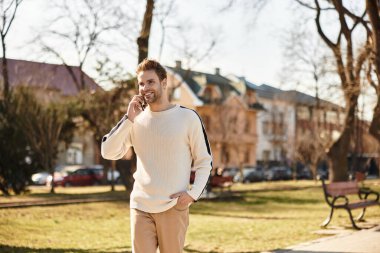 Neşeli bir adam, güneşli bir parkta yürürken telefonuyla sohbet ediyor..