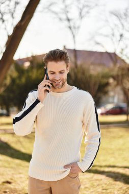 Şık bir genç adam güneşli bir parkta yürürken, telefonda konuşurken gülümsüyor..