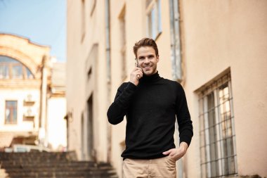 Şık bir adam, güneşli bir sokakta güvenle yürüyor, telefonla konuşuyor ve gülümsüyor..