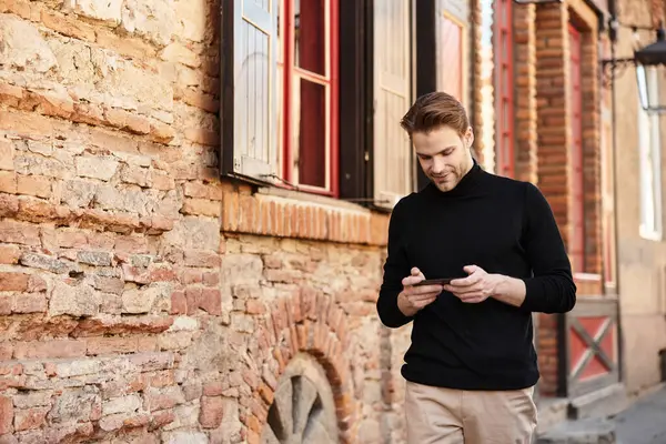Mobil cihazına odaklanmış şık bir adam tarihi bir sokakta gelişigüzel yürüyor..