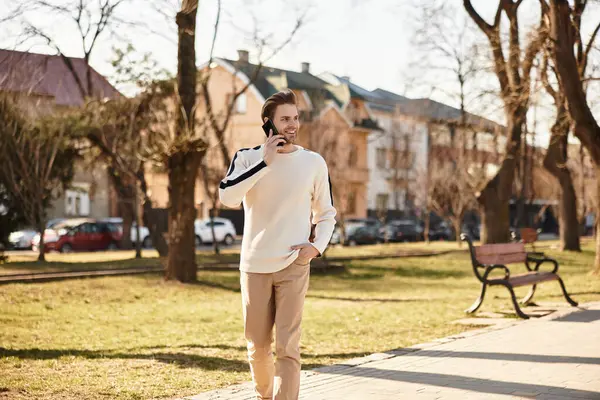 Çekici bir adam güneşli bir parkta rahatça yürüyor, cep telefonuyla sohbet ediyor..