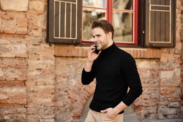 Um homem elegante caminha confiantemente ao longo de uma rua histórica, conversando em seu telefone com um sorriso. — Fotografia de Stock