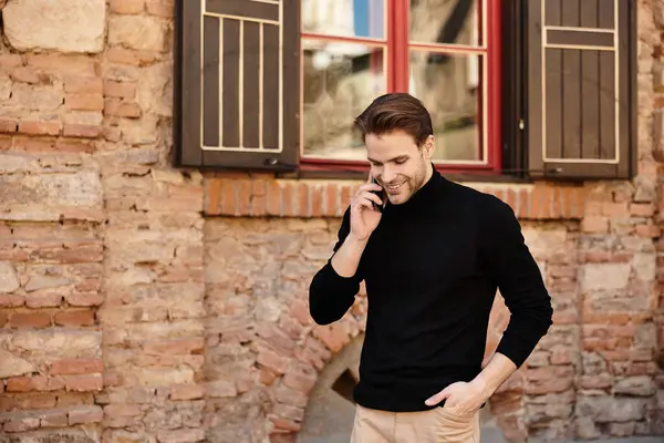Un bell'uomo cammina lungo un rustico muro di mattoni, parlando al telefono con un'espressione allegra. — Foto stock