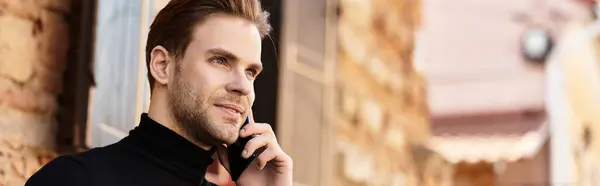 A confident man walks along a quaint street, engaged in a phone conversation under a soft sun. — Stock Photo