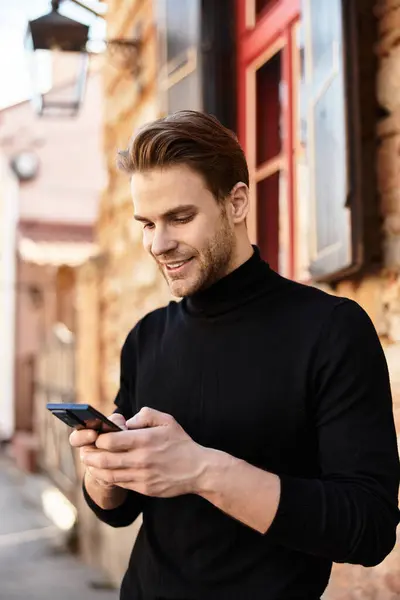 Um jovem elegante passeia por uma rua pitoresca, imerso em seu smartphone enquanto sorri. — Fotografia de Stock