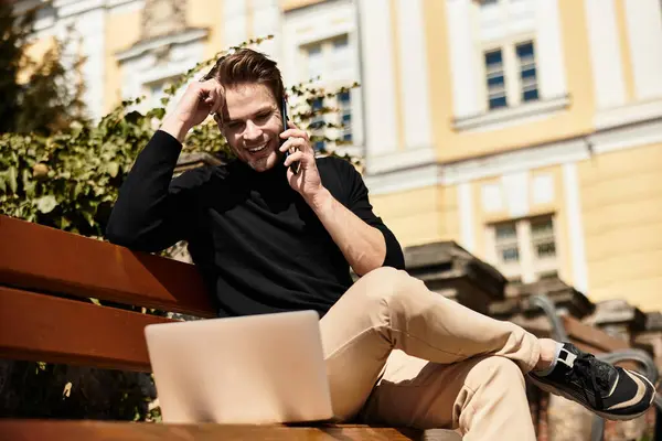 A stylish man sits on a bench, talking on the phone and smiling while working on his laptop. — Stock Photo