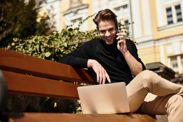 A stylish man with a confident smile sits on a bench, chatting on his phone while using a laptop. — Stock Photo