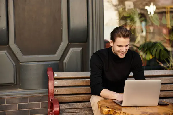 A handsome man smiles at a wooden table while working on his laptop outdoors. — Stock Photo