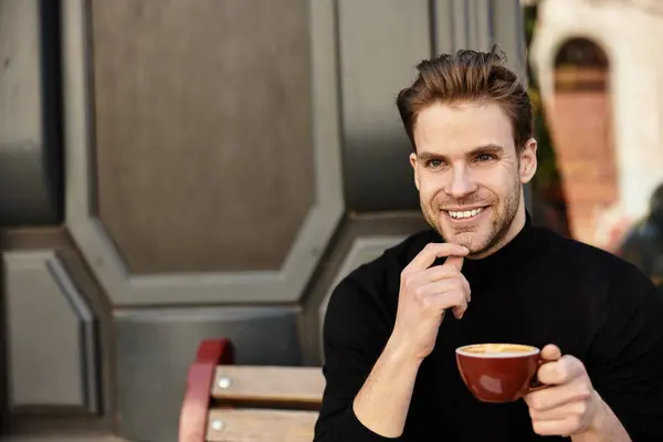A handsome man enjoys coffee at a cozy cafe, smiling at passersby on a relaxing afternoon. — Stock Photo