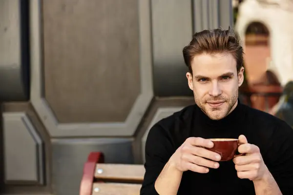 A charming young man sits on a bench, savoring a warm beverage in a lively city setting. — Stock Photo
