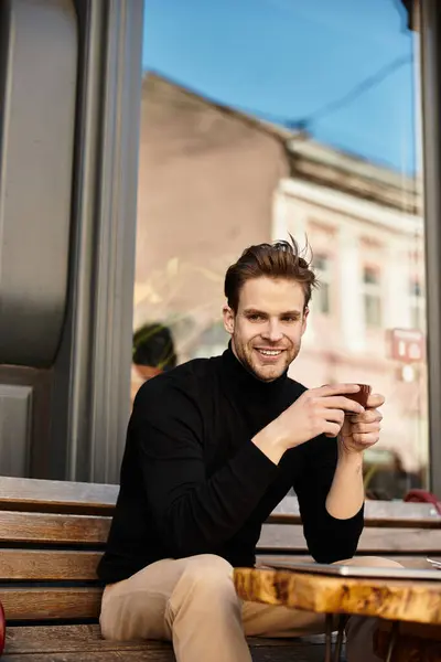 A charming man sits on a bench, smiling gently while sipping coffee, basking in warm sunlight. — Stock Photo