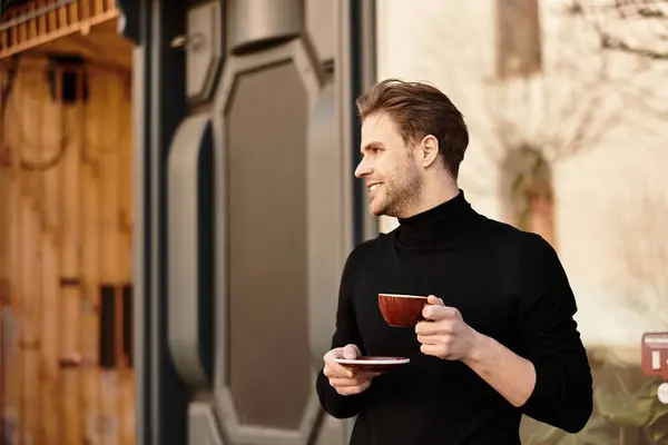 Man with a cheerful smile holds a cup and saucer while walking through a quaint outdoor setting. — Stock Photo