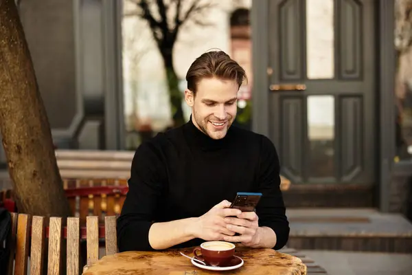 Ein gutaussehender Mann lächelt in sein Telefon, während er draußen in der Nähe eines gemütlichen Cafés Kaffee genießt. — Stockfoto
