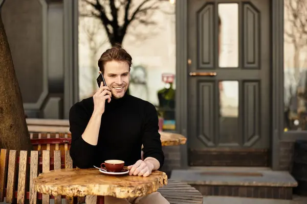 Handsome man sits at a wooden table, smiling and talking on the phone, with coffee beside him. — Stock Photo