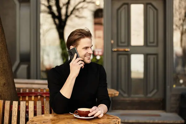 Handsome man walks and talks on his phone, savoring coffee outside in a lively urban cafe. — Stock Photo