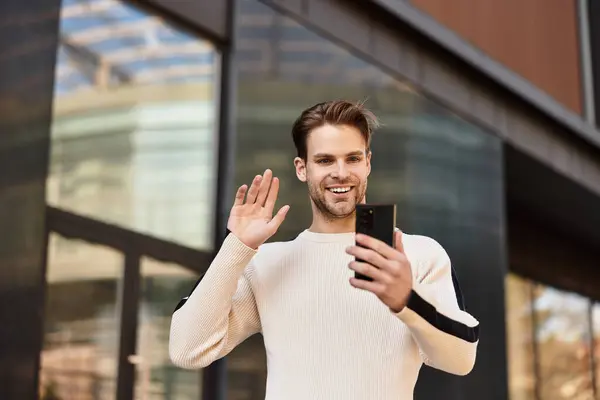 A stylish man smiles and waves while walking through a modern city, using his smartphone. — Stock Photo
