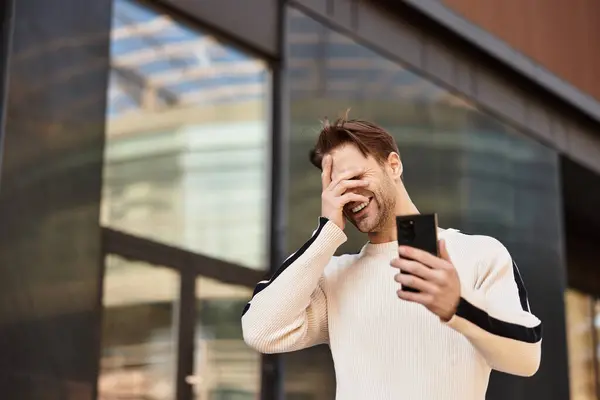 A charming man strolls through the city, laughing joyfully while looking at his phone. — Stock Photo