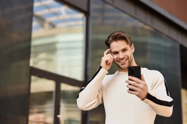 A stylish man strolls through a modern cityscape, engaged with his smartphone and smiling brightly. — Stock Photo