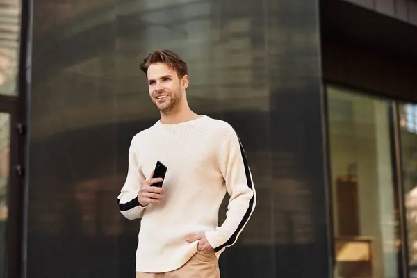 Hombre confiado pasea por la calle, centrado en su teléfono inteligente por edificios modernos. - foto de stock