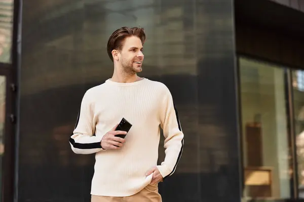 A stylish man with a bright smile walks the city, smartphone in hand, looking relaxed. — Stock Photo