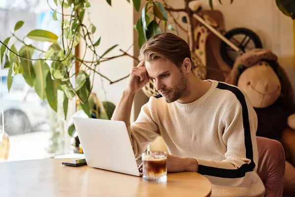 A handsome man sits at a wooden table, focused on his laptop while enjoying a drink. — Stock Photo