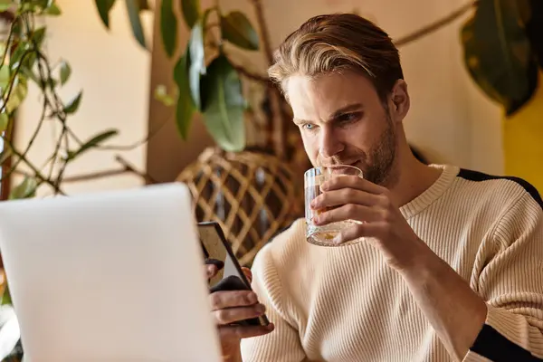 Un hombre guapo bebe mientras se conecta con su teléfono y su computadora portátil en un ambiente cálido y acogedor. - foto de stock