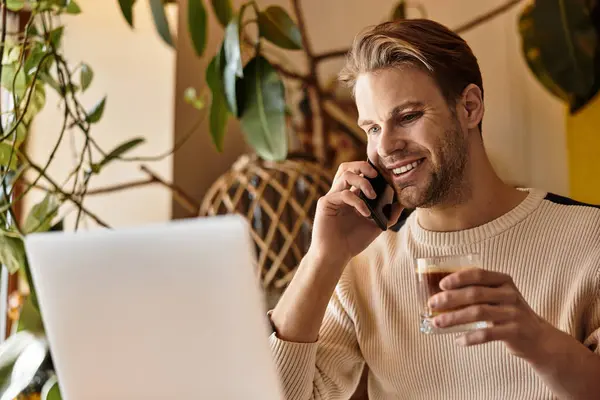 A stylish man happily engages in a phone call while sipping coffee, focused on his laptop. — Stock Photo