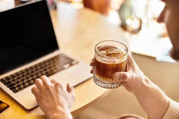 A handsome man relaxes at a table with a drink while focusing on his laptop in a lively area. — Stock Photo