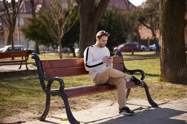 Um homem bem vestido caminha vagarosamente por um parque tranquilo, absorvido em seu smartphone. — Fotografia de Stock
