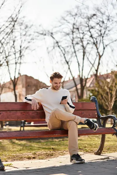 Enjoying the warmth of the sun, a stylish man checks his phone while sitting on a bench in a park. — Stock Photo