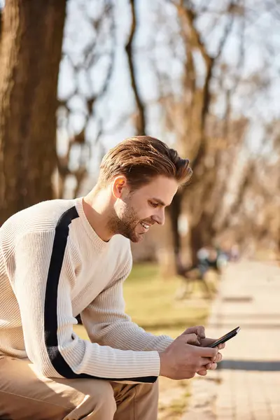 A stylish man strolls through a beautiful park, smiling as he checks messages on his phone. — Stock Photo