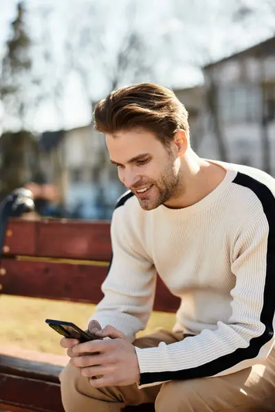 A stylish man sits on a bench outdoors, absorbed in his smartphone, surrounded by nature. — Stock Photo