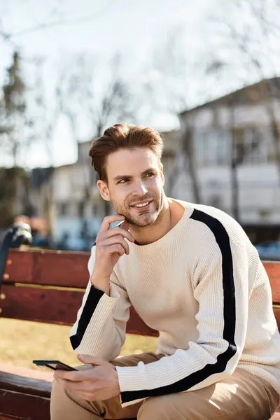 Handsome man casually strolls through a lively park, smiling and engaging with his phone. — Stock Photo