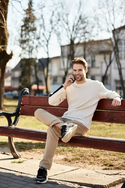 A young man smiles on a park bench, enjoying a phone call in the sunshine. — Stock Photo