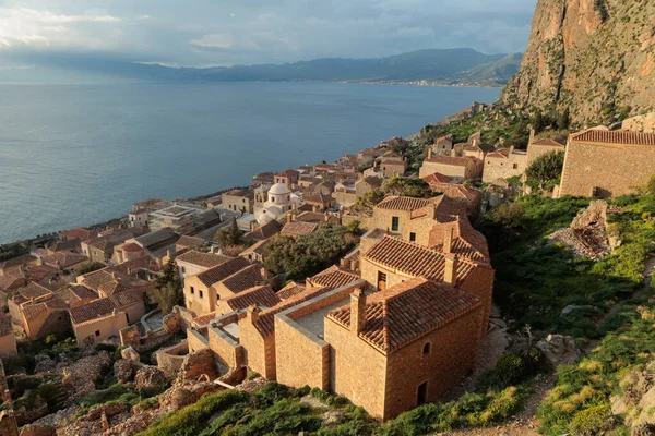 View of fortified medieval castle village Monemvasia with Aegean sea and Lakonia coast as background. Stone houses with dusty pink terracotta tiled roofs and blue sea. Antique buildings under soft