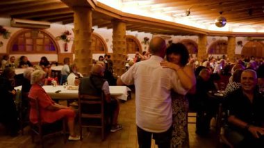 Gülümseyen yaşlı bir çift sıcak bir İspanyol restoranında canlı müzik eşliğinde neşeyle dans ederken diğerleri yemeğin tadını çıkarıyor. Sıcak atmosfer, otantik eğlence, mutlu an