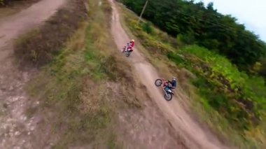 Exciting FPV drone chase of two motocross riders competing on a forest dirt track. Riders jump ramps, race at high speed, and showcase extreme motocross action from a cinematic aerial perspective