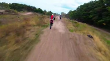 Exciting FPV drone chase of two motocross riders competing on a forest dirt track. Riders jump ramps, race at high speed, and showcase extreme motocross action from a cinematic aerial perspective