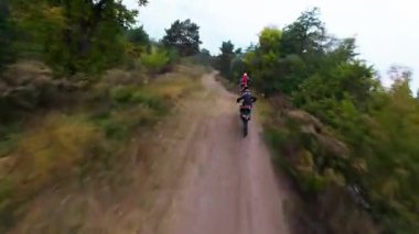 Exciting FPV drone chase of two motocross riders competing on a forest dirt track. Riders jump ramps, race at high speed, and showcase extreme motocross action from a cinematic aerial perspective
