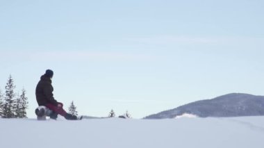 Tired alpinist takes a break to catch a breath and think at the scenic valley covered in snow. Camera pans at frozen Christmas trees