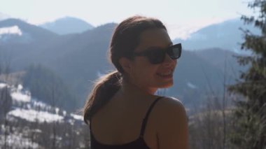 Gözlüklü kız kameraya gülümser ve ilkbaharda sakin dağ sıralarını gözlemlemek için arkasını döner.