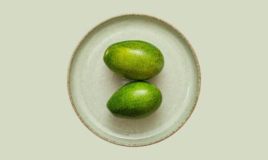Avocado İkili. İki mükemmel olgun avokado, yumuşak pastel yeşil bir zemine karşı sunulan seramik bir tabakta oturur. Meyvenin pürüzsüz ve parlak dokusunu vurgularlar. Üst görünüm.