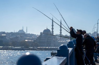 Balıkçı, Galata Köprüsü 'nde balıkçılık yapıyor. Her gün kalabalık bir köprü kenarı. Arka planda Yeni Cami Camisi var.