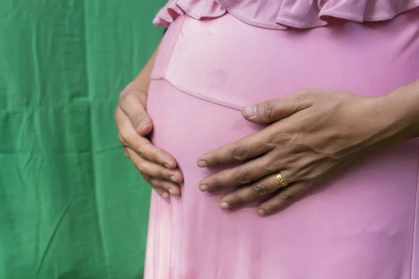 Pembe elbiseli kadın hamile karnına dokunuyor. Yeşil arka planda hamile anne göbeği.