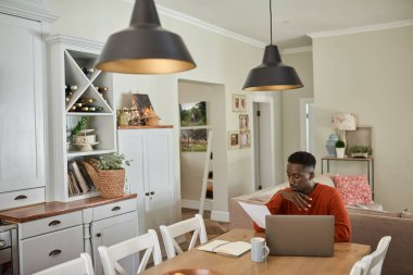 Young male entrepreneur reading paperwork and using a laptop while working remotely from home at a dining room table