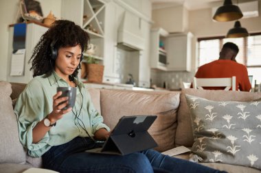Young African woman wearing headphones and using a tablet on a sofa with her husband working at a kitchen table behind her