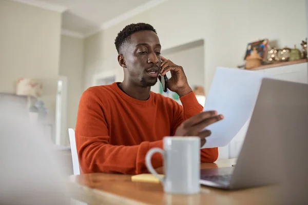 Young African male entrepreneur talking on his phone and going over paperwork while working remotely from home on a laptop