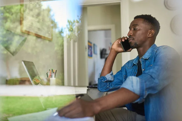 Young African male entrepreneur talking on a phone and working on a laptop at a desk while sitting inside of his home office