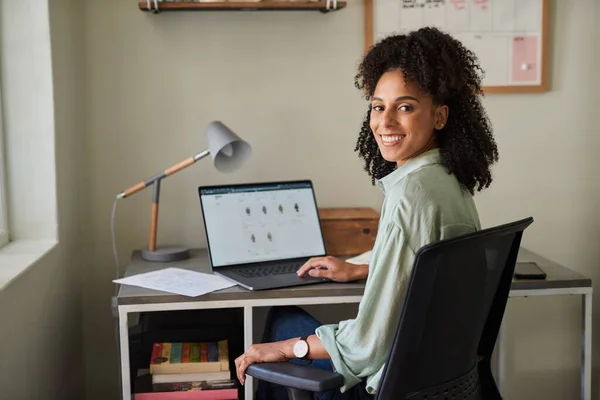 Portrait of a smiling young African female entrepreneur sitting at a desk in her home office and working online with a laptop